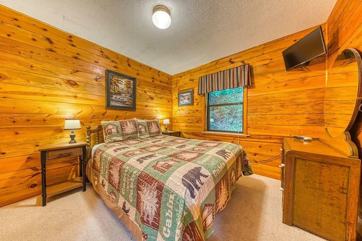 A warmly lit bedroom with wooden walls, a bed covered in a patterned quilt, a window with a view to the outdoors, a bedside lamp, and a TV mounted on the wall. The space feels inviting and comfortable, blending natural textures with cozy accents.