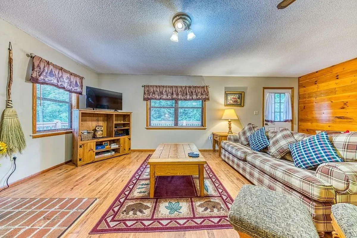 The scene highlights a comfortable plaid sofa paired with a wooden coffee table and a TV stand adorned with decorative items. The space is well-lit and arranged for group entertainment, ideal for movie nights or playing board games.