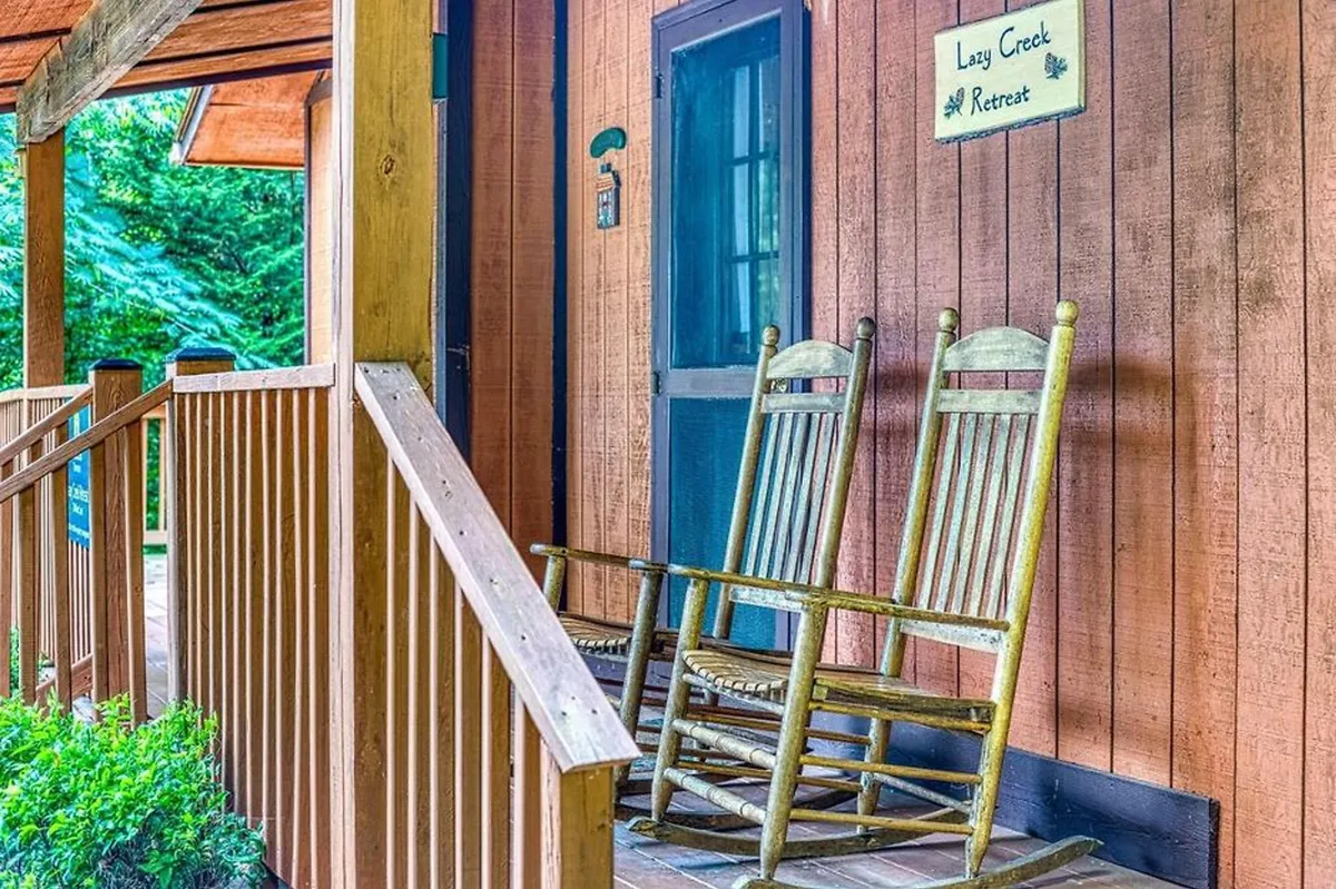This exterior image shows a charming, covered front porch with classic wooden rocking chairs and a wooden railing. A sign reads 'Lazy Creek Retreat,' reinforcing the sense of place. The porch is surrounded by lush greenery, and the setup invites quiet moments while enjoying the fresh mountain air.