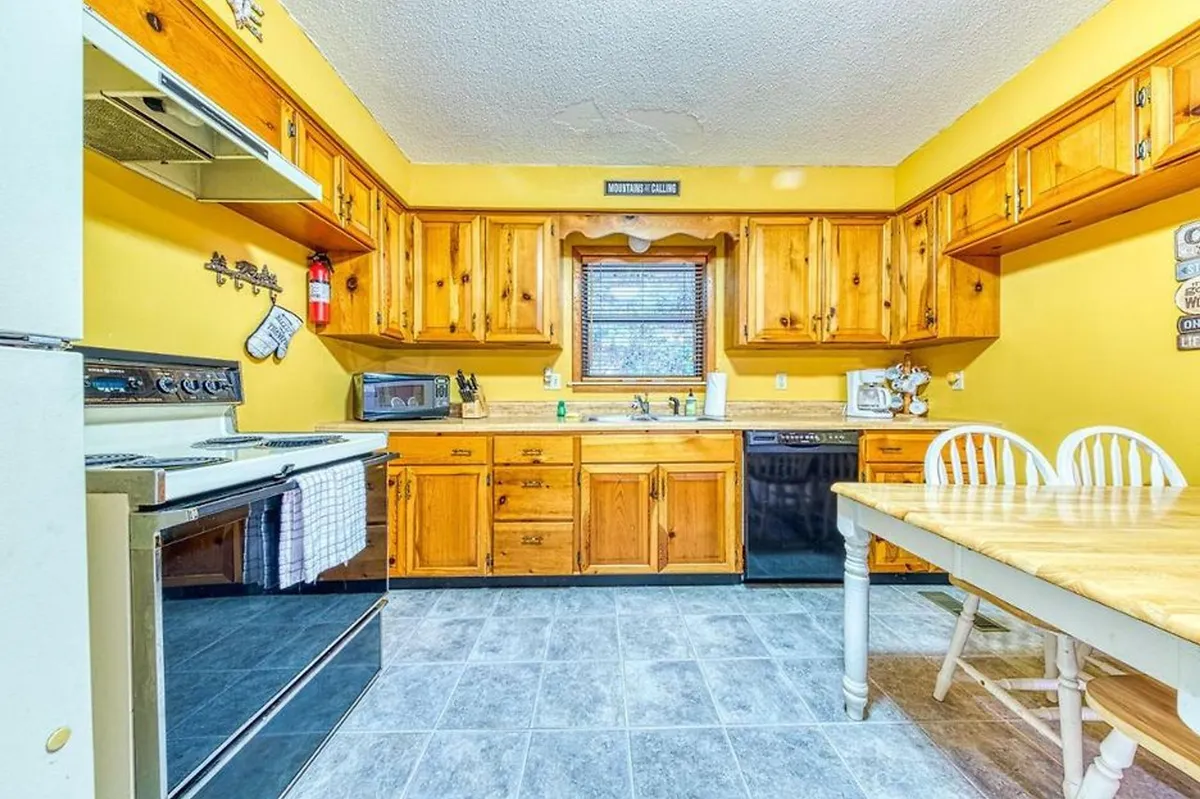 This image captures a wide view of the kitchen and dining area, showcasing wooden cabinetry, a kitchen island, and a dining table set by a window with blinds. The warm tones and inviting arrangement highlight the space's functionality and comfort for group meals and gatherings.