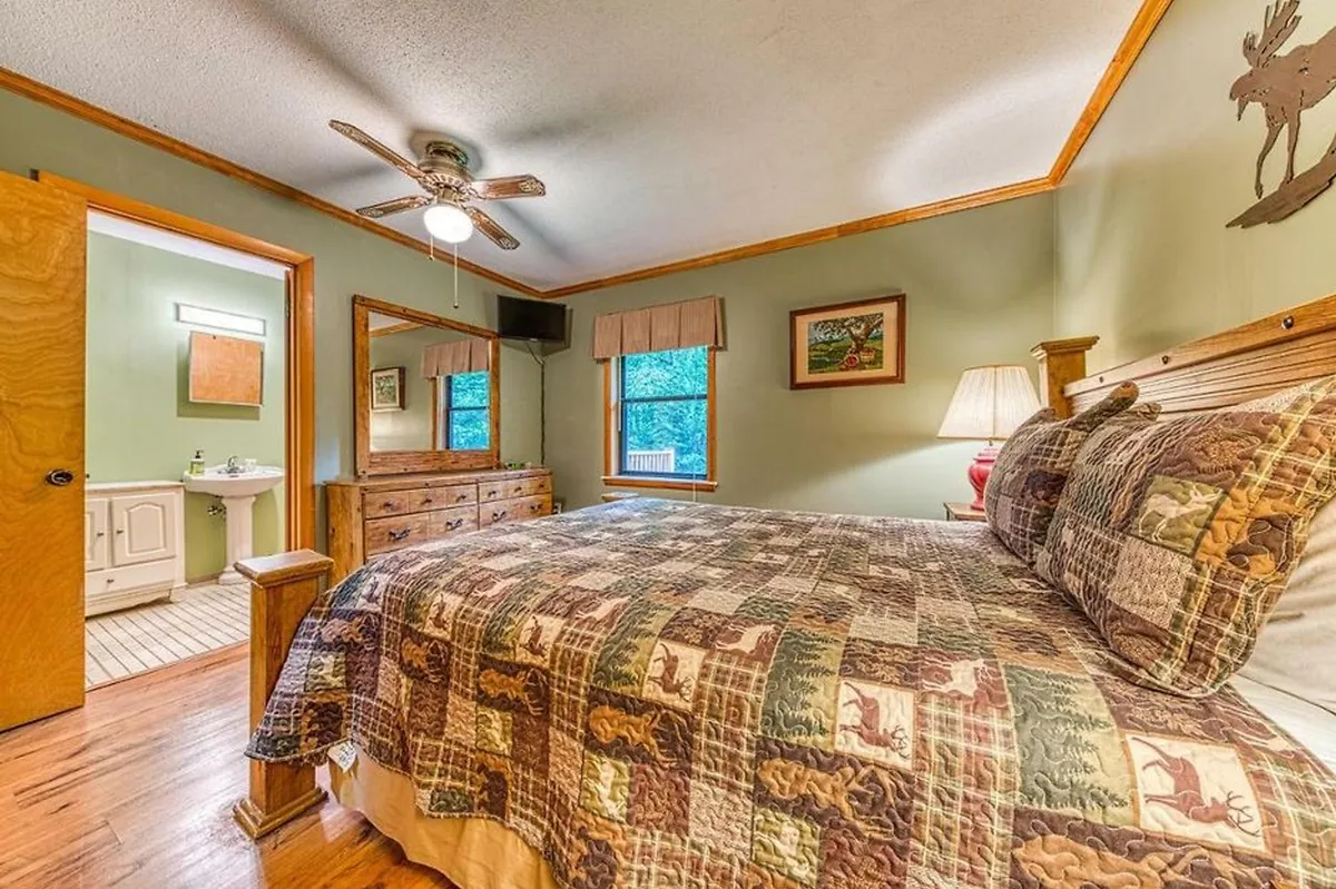 A warmly lit bedroom with a patterned quilt on the bed, wooden dresser, and curtained windows that allow sunlight to flood the room. The scene exudes comfort and encourages slow, restful mornings.