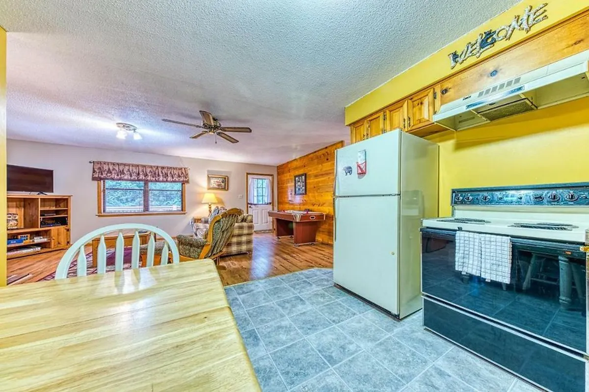 A broad interior view that seamlessly connects the kitchen area, dining table, and adjacent living space, with natural light streaming through the window. The setup suggests a multi-functional, open-plan area ideal for cooking, dining, and relaxing together.