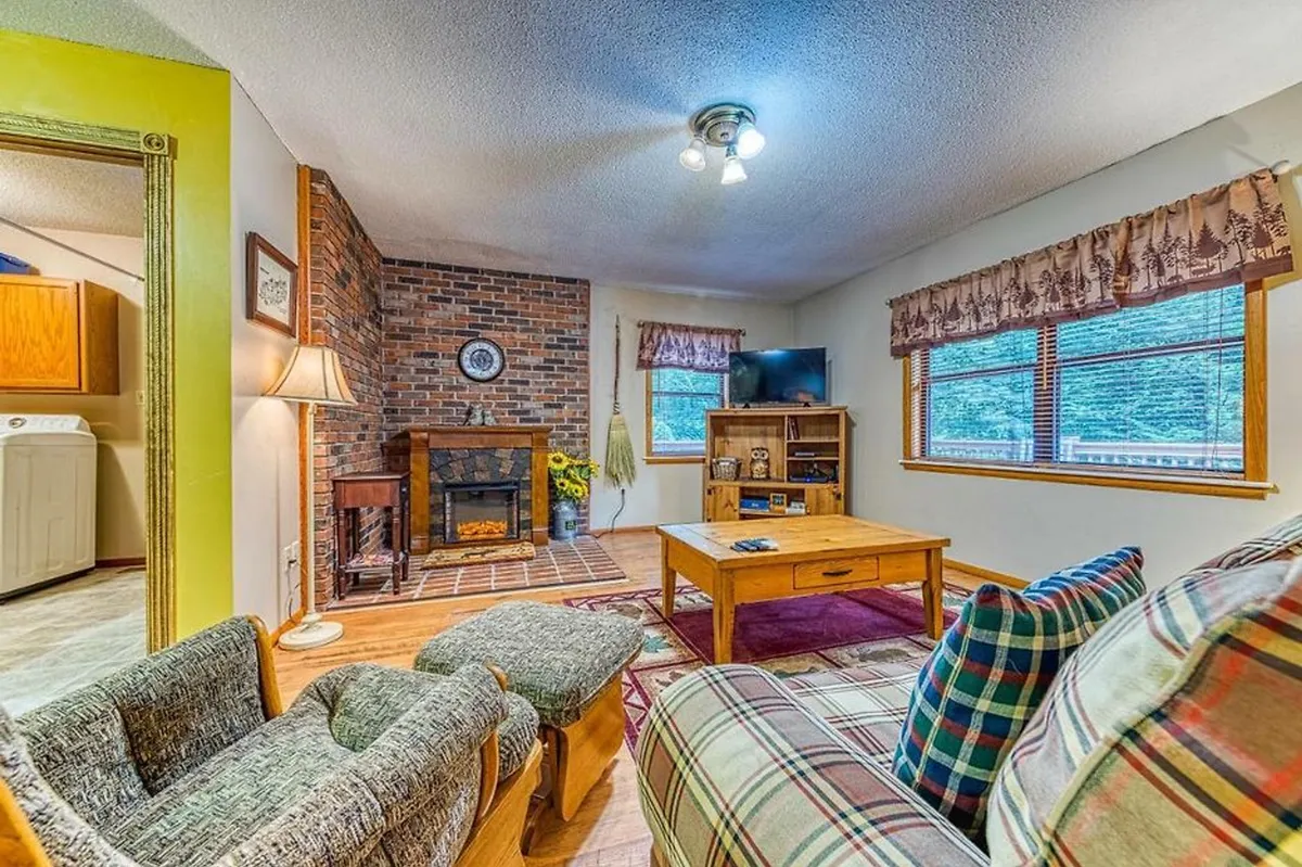 This image showcases a welcoming living area with a brick fireplace, a plaid sofa, a wooden coffee table, and a television. Warm wood paneling and soft lighting create an inviting atmosphere perfect for relaxation and social gatherings. The TV, fireplace, and comfortable furnishings encapsulate the essence of cozy mountain living.