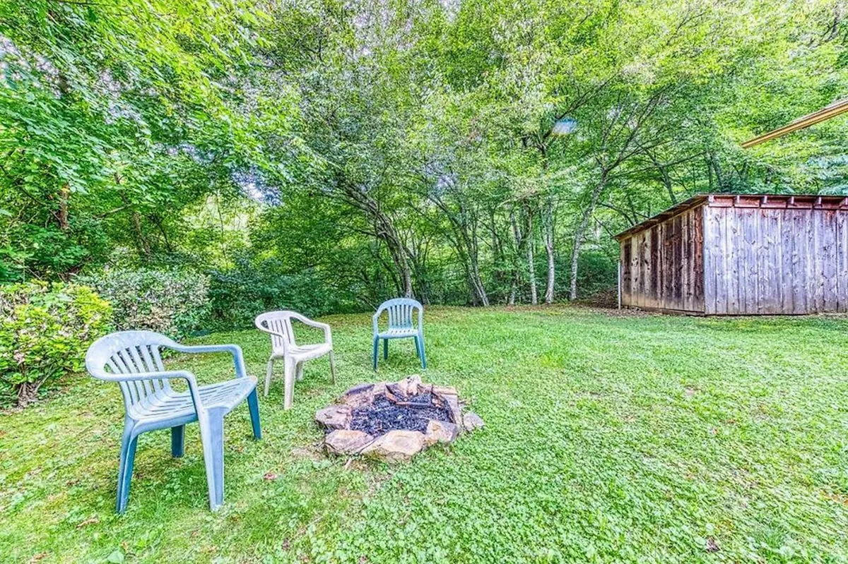 Nestled in the yard, a cozy fire pit is encircled by comfortable lawn chairs. The scene is framed by leafy trees and open space, providing the perfect spot for evening gatherings, storytelling, or stargazing by firelight.
